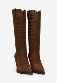 Botas de ante marrón hasta la rodilla con punteras afiladas, acentos en forma de ondas y cremallera lateral. Textura suave y tacón en cuña afilado.