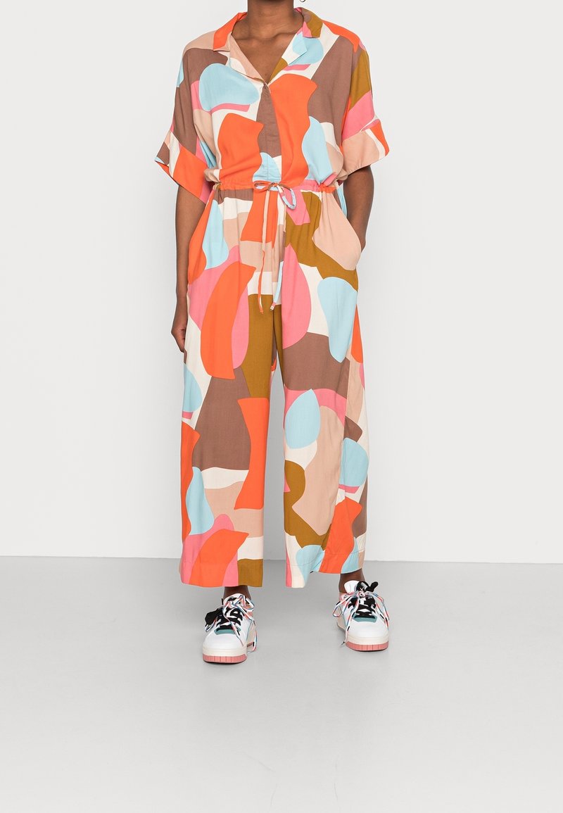 Personne portant une combinaison aux motifs abstraits colorés avec manches courtes et des baskets blanches à lacets multicolores.