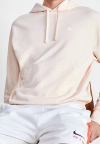 Ljusrosa hoodie gjord av mjukt tyg, med en ficka fram och en liten vit Nike-logga på bröstet. Parat med vita shorts.