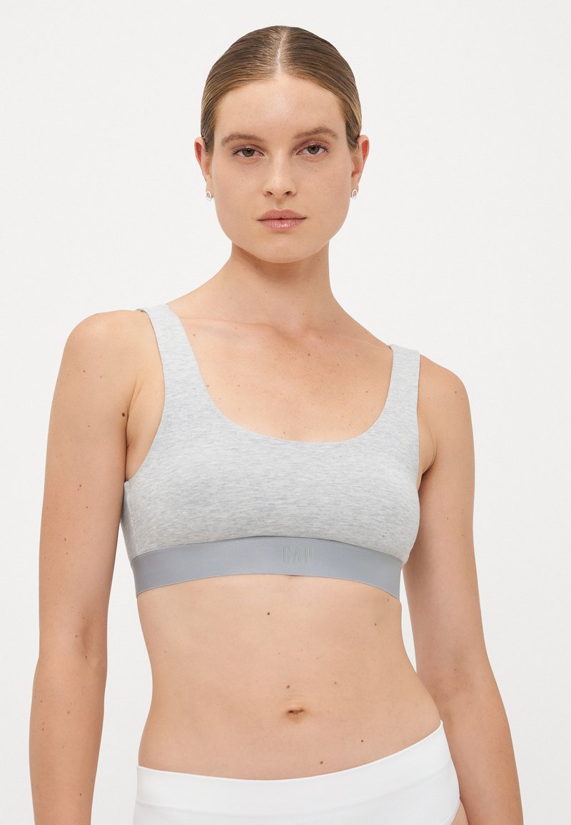 Woman with slicked-back hair wearing a light gray sports bra and white bottoms against a plain white background.
