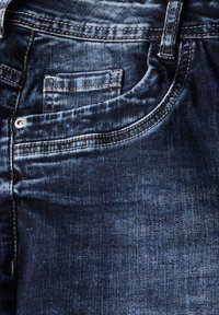 Tummansiniset farkut, joissa on etutasku, näkyvät saumat ja hieman kulunut pinta, mikä korostaa klassista muotoilua.