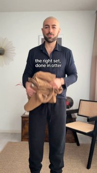 Άνδρας με ξυρισμένο κεφάλι και γένια κρατά ένα διπλωμένο καφέ σακάκι, όρθιος σε ένα δωμάτιο με καρέκλα, γραφείο και διακόσμηση τοίχου με σχέδιο ηλιακών ακτίνων. Το κείμενο γράφει «το σωστό σετ, φτιαγμένο σε κεντρίδα».