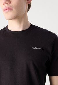 Fekete pamut póló kerek nyakkivágással. Kicsi, fehér "Calvin Klein" logó található a bal mellen. Sima textúrájú, klasszikus szabású.