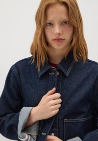 Jeune femme aux cheveux roux portant une veste en denim bleu foncé, tenant le devant de la veste d'une main, devant un fond uni.