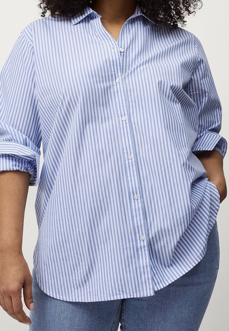 Femme portant une chemise rayée bleu et blanc à boutons avec les manches retroussées et un jean bleu, main dans la poche.