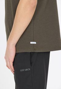 Oliivinvihreä puuvillapaita, jossa on suora leikkaus ja lyhyet hihat; helmassa on pieni valkoinen etiketti, jossa lukee "NEW BAD HABITS".