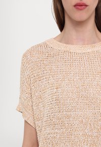 Haut en maille de couleur pêche claire et crème avec une coupe ample. Doté d'un col large et de manches courtes. Détails en texture à trame ouverte présents.