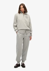 Sudadera gris con cremallera y logotipo bordado de "Superdry Athletics", combinada con joggers grises y una camisa a rayas de capas. Material suave, ajuste relajado.