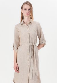 Robe chemise en lin beige avec une fermeture à boutons, des poches latérales, des manches retroussées et une ceinture assortie à la taille. Tissu texturé.
