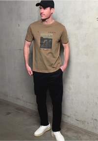 Brun bomull t-shirt med ett tryckt motiv av träd, matchad med svarta byxor och vita sneakers, mot en betongvägg.