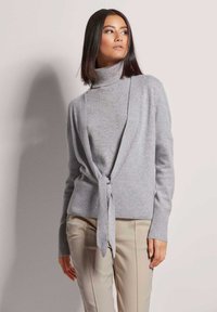 Lichtgrijze gebreide trui met een col en een gestrikte sluiting aan de voorkant. Heeft ribbelmanchetten en een ribbelzoom, gecombineerd met beige slim-fit pantalons.