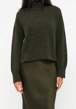 Femme portant un pull à col roulé vert foncé à manches longues et une jupe ajustée assortie sur un fond uni.