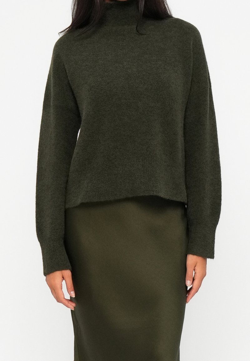 Femme portant un pull à col roulé vert foncé à manches longues et une jupe ajustée assortie sur un fond uni.