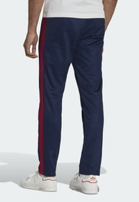 Mörkblå träningsoveraller med röda ränder längs sidorna, tillverkade av ett slätt material. Kombineras med vita sneakers med detaljer och logotyp.