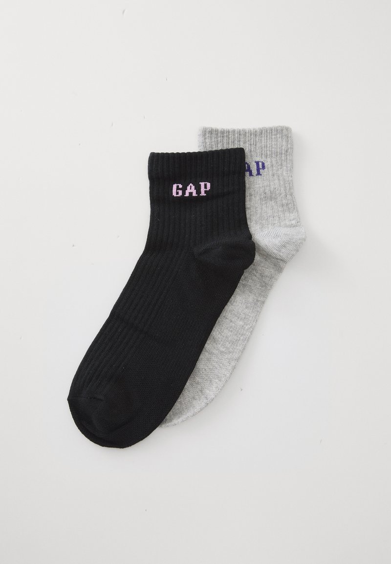 Une paire de chaussettes côtelées arrivant à la cheville, l'une noire avec le texte "GAP" en rose et l'autre gris clair avec le texte "GAP" en violet, sur un fond blanc.