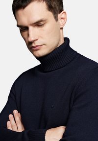 Ciemnoniebieski sweter typu golf z prążkowanym kołnierzem i mankietami. Dzianinowa faktura, dopasowany krój, z małym akcentem logo na piersi.