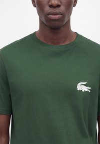 Personne à la peau foncée portant un t-shirt vert foncé avec un petit logo blanc de crocodile sur le côté gauche de la poitrine.