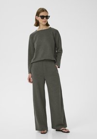 Felpa grigia oversize e pantaloni a gamba larga realizzati in morbido materiale, abbinati a occhiali da sole neri e ciabatte.