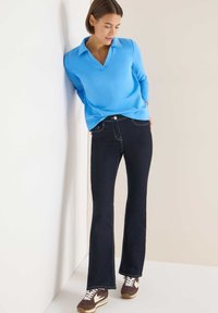 Lyseblå langærmet top med V-hals og krave kombineret med mørke, vide jeans. Brune sneakers fuldender outfittet.