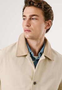Joven con ojos azules que lleva una chaqueta beige sobre una camisa de denim azul, mirando hacia un lado contra un fondo liso.
