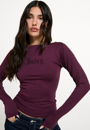 Žena s dlhými tmavými vlasmi, ktorá nosí priliehavé fialové tričko s dlhým rukávom s nápisom "Juicy" na hrudi a tmavé rifle.