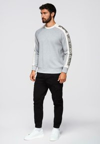 Sudadera gris con mangas raglán, con acentos blancos y texto negro. Combinada con pantalones cargo negros y zapatillas blancas.