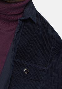 Camisa de pana marine con un bolsillo frontal, combinada con un suéter de cuello cisne burdeos ajustado. Presenta un detalle de botón en el bolsillo.