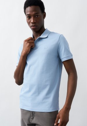 Νεαρός άνδρας που φοράει ανοιχτό γαλάζιο κοντομάνικο πόλο μπλουζάκι και γκρι παντελόνια, ρυθμίζοντας το γιακά του πουκαμίσου, στέκεται μπροστά σε λευκό απλό φόντο.