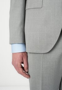 Light gray suit with a textured pattern; a blue shirt cuff is visible. The suit jacket has a plain pocket and a tailored fit.
