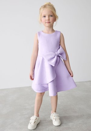 Lilla vabaõhukuue, millel on pael, tekstuuriga mustriga tähed ja paelad, laienev siluett, koos valgete sneakersitega, millel on kleepribad.
