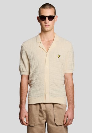 Uomo che indossa una camicia beige a maniche corte con bottoni e texture, con logo di uccello giallo, pantaloni color camoscio e occhiali da sole neri su sfondo grigio chiaro.