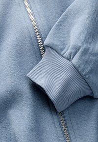 Light blue zip-up jacket featuring ribbed cuffs and a textured fabric. The zipper runs down the front, showcasing a clean, modern design.
