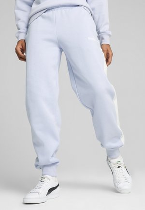 Person iført lyseblå Puma joggingbukser med hvide sidelister og hvide Puma sneakers står mod en ensfarvet baggrund.
