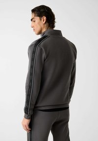 Giacca da allenamento grigia con zip, collo alto, tessuto strutturato e strisce nere decorative con logo lungo le maniche.