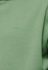 Sudadera verde claro con una tela suave y texturizada. Presenta un logo en relieve "PECADOR" en un color a juego en la parte delantera.