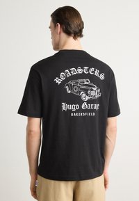 Black cotton t-shirt featuring a white graphic print of a vintage car and text saying "ROADSTERS" and "Hugo Garage BAKERSFIELD" on the back.