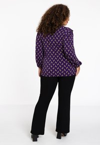 Lila Bluse mit geometrischem Muster, langen Ärmeln und einem runden Ausschnitt, kombiniert mit schwarzen Schlaghosen und schwarzen Blockabsatzschuhen.