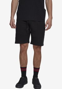 Shorts de sport noirs avec une taille élastiquée et cordon de serrage, arrivant au genou, avec poches latérales et une texture lisse. Associés à des baskets noires et des chaussettes rayées.