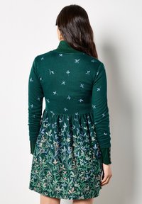 Abito verde con maniche lunghe, caratterizzato da un motivo di uccelli sulla parte superiore e un design floreale sulla gonna. Tessuto morbido con vita arricciata.
