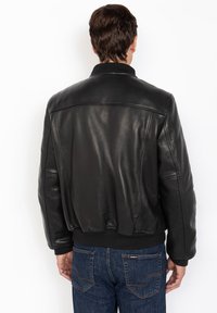 Uomo con giacca di pelle nera e jeans blu, di spalle, in piedi contro uno sfondo bianco.