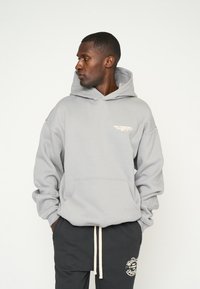Felpa grigia con tasca a marsupio, cappuccio con coulisse e logo ricamato in beige chiaro sul petto. Tessuto morbido e vestibilità comoda.