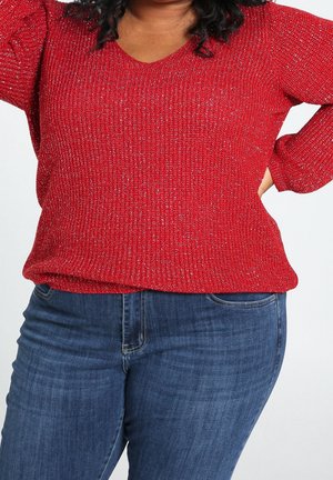 Personne portant un pull en tricot rouge et un jean bleu, une main posée sur la hanche, sur un fond clair uni.