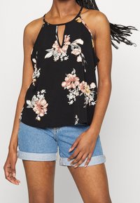 Débardeur noir à motifs floraux avec un détail à ouverture en forme de goutte, présentant des motifs de fleurs roses et blanches. Porté avec un short en jean bleu, offrant une coupe décontractée.