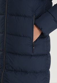 Marineblå puffjacka med kviltad struktur, sidoficka med dragkedja och en tät passform med en smidig materialfinish.