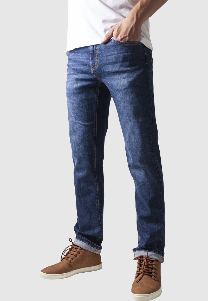 Persona con jeans blu risvoltati e scarpe marroni stringate, con una mano in tasca, su sfondo uniforme.