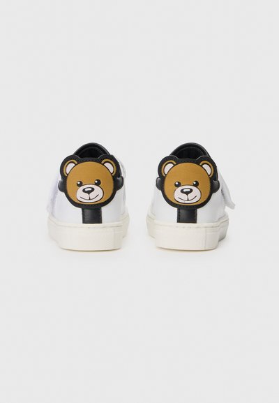 White sneakers with black and brown bear face patches on the heel, featuring a smooth texture and a flat rubber sole.