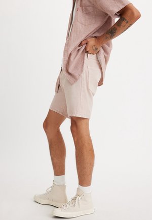 Homme portant un short rose pâle, une chemise à manches courtes assortie, des chaussettes blanches et des baskets montantes beiges, debout avec une main dans la poche.