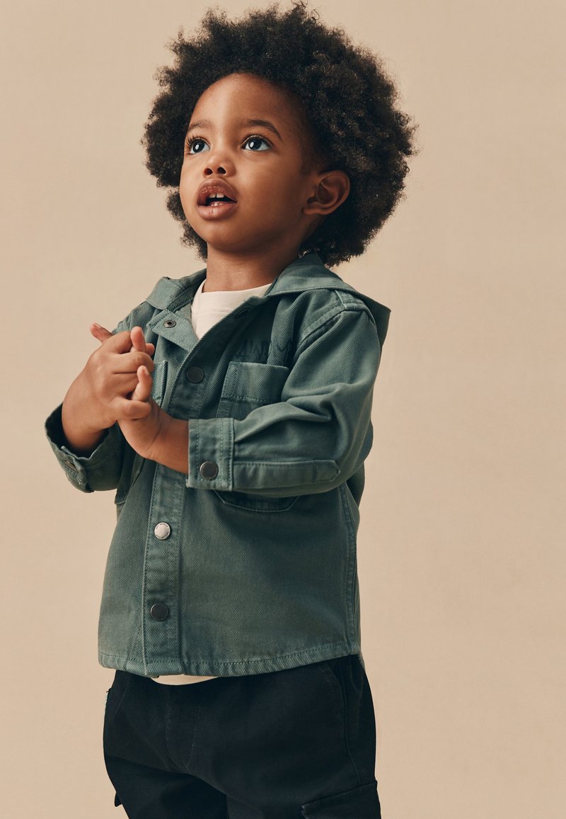 Jeune enfant aux cheveux bouclés portant une veste verte, regardant vers le haut, les mains jointes, sur un fond beige uni.