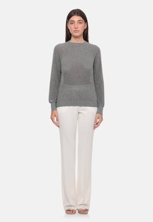 Donna in piedi di fronte, indossa un maglione grigio a maniche lunghe e pantaloni bianchi a gamba dritta con sandali aperti su sfondo neutro.