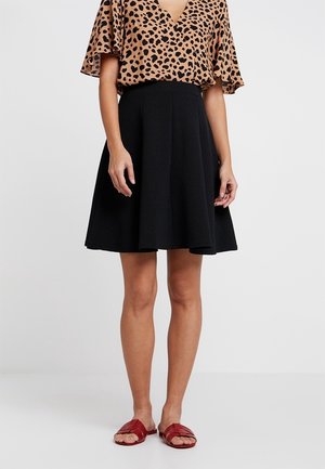 Jupe noire évasée avec des plis, fabriquée à partir d'un tissu texturé. Assortie à un haut en imprimé léopard brun et des sandales à glissière rouges.